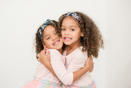 Two young girls with curly hair wearing AYOCATHY elastic back turban style silk headband. Girls are wearing pink dresses and hugging each other.